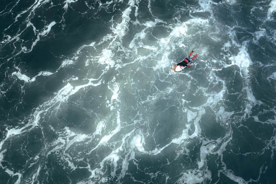 Ein Surfer paddelt auf einem Surfbrett liegend auf dem offenen Meer über das Wasser, um ihn herum Schaumkronen von gebrochenen Wellen.