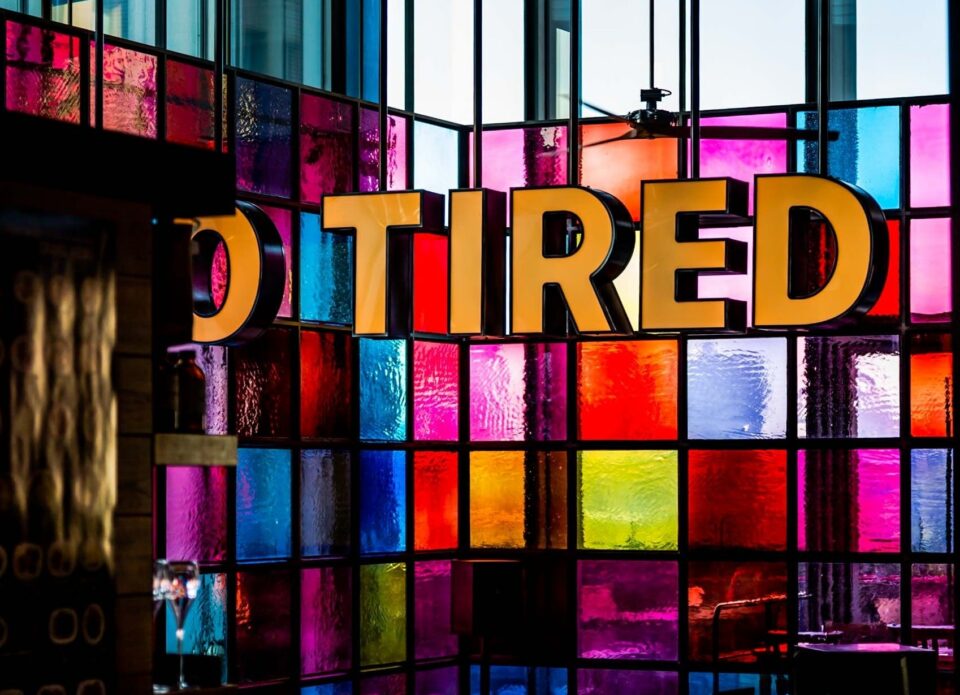 Schriftzug „TIRED“ vor einer bunten Glaswand.