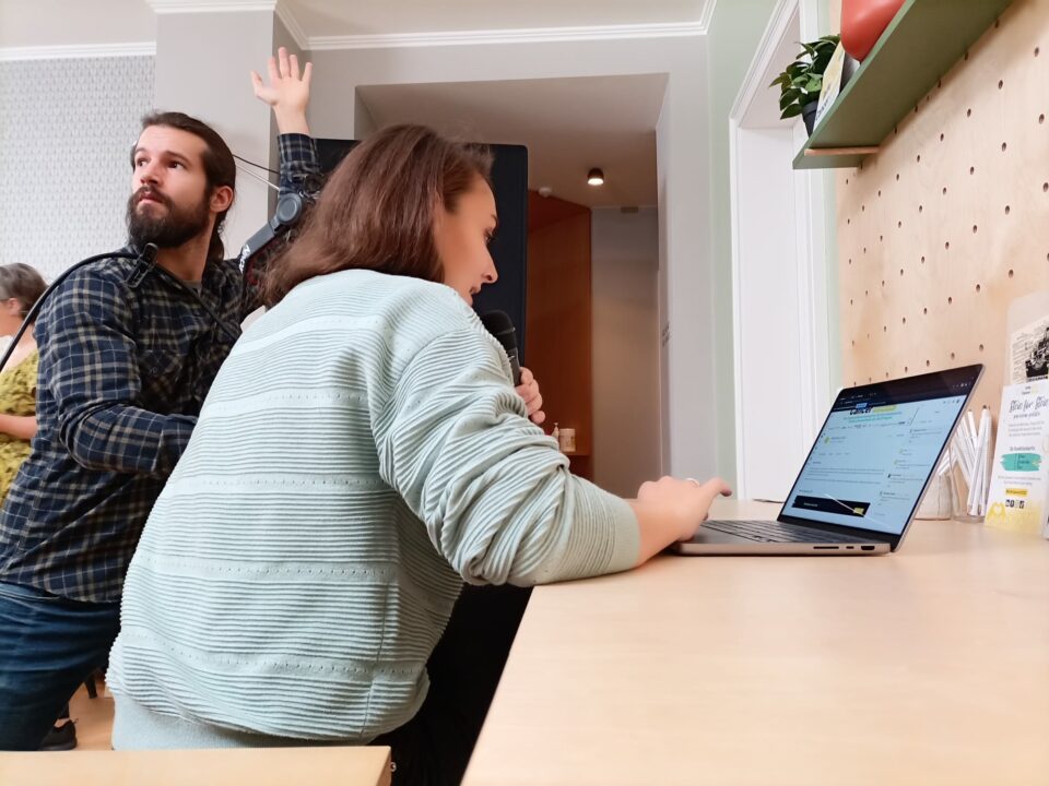 Lena moderiert vor dem Laptop, während der Kameramann die Hand hebt.