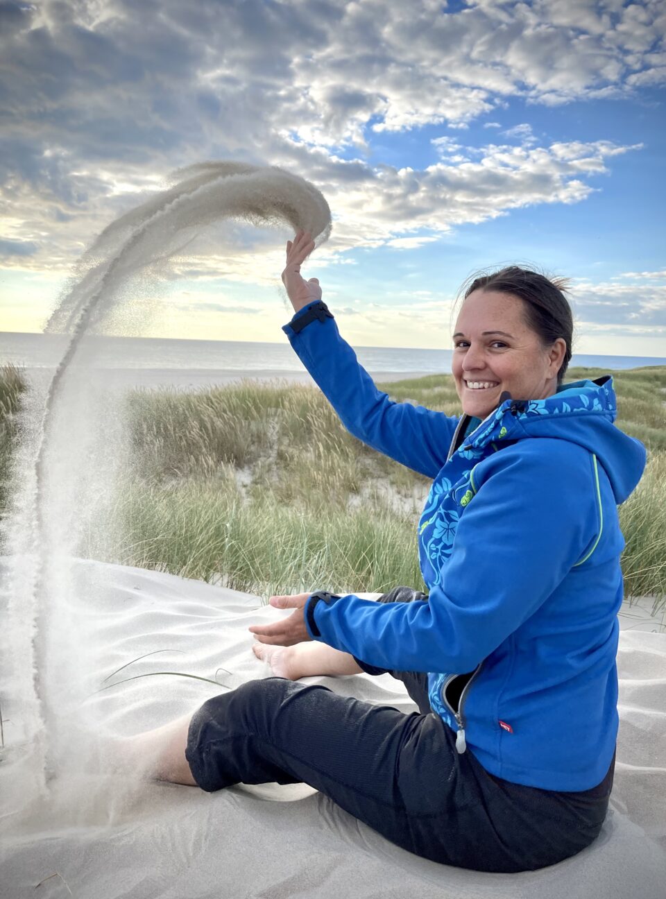 Frau am Strand wirft Sand in die Luft und lächelt dabei in die Kamera