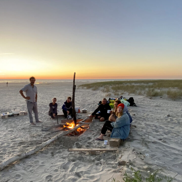 Das Kurvenkratzer-Team sitzt bei Sonnenuntergang an einem Lagerfeuer am Strand.