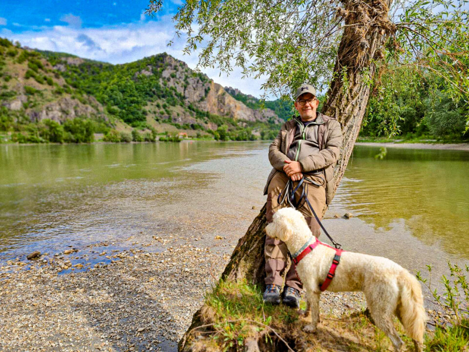 Mann mit Hund lehnt an Baumstamm am Fluss